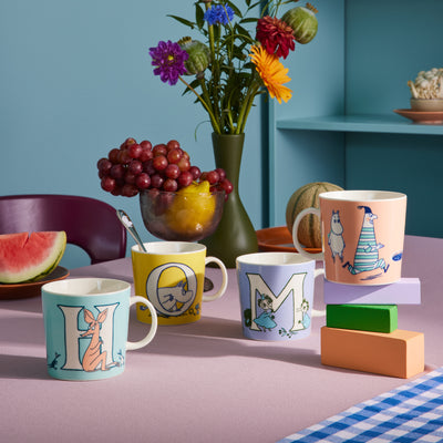 Colorful mugs with letter designs on a table with fruits and flowers.