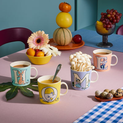 Table setting with colorful mugs, snacks, and fruits on a checkered tablecloth.