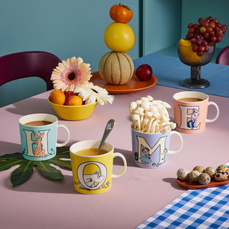 Table setting with colorful mugs, snacks, and fruits on a checkered tablecloth.