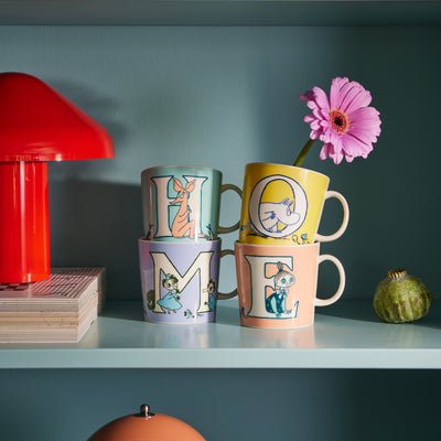 Four colorful mugs with cartoon designs on a shelf against a teal wall.