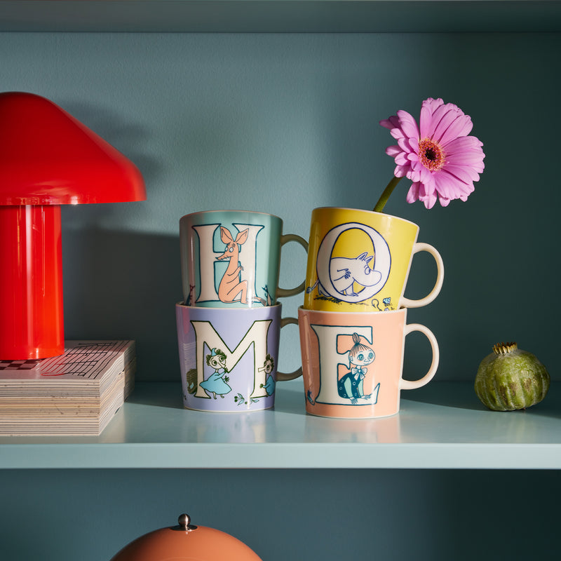 Four colorful mugs with cartoon designs on a shelf against a teal wall.