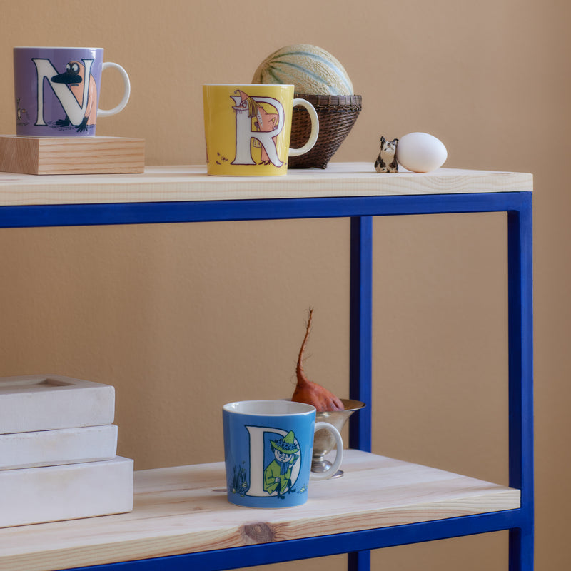 Colorful mugs with letter designs on a wooden shelf against a beige wall.