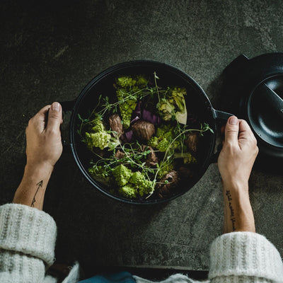 Person holding a black frying pan with green vegetables on a dark surface