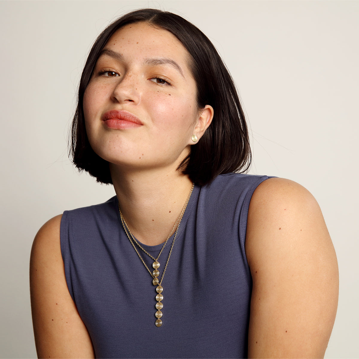 Woman wearing a purple sleeveless top and gold necklace against a plain background