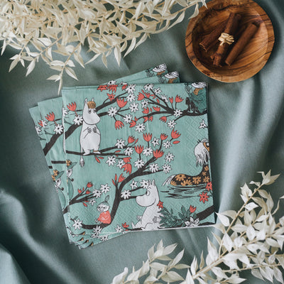 Two Moomin-themed napkins on a green fabric background with a wooden bowl of cinnamon sticks.