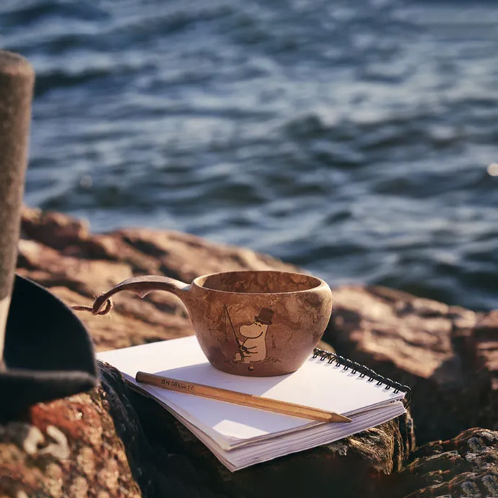 Kupilka Moominpappa Classic Cup sitting on notebook near rocky edge of water
