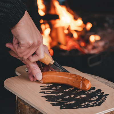 Slicing sausage on Muurla Pine Cone/Birch Leaf Chop & Serve Board by fire